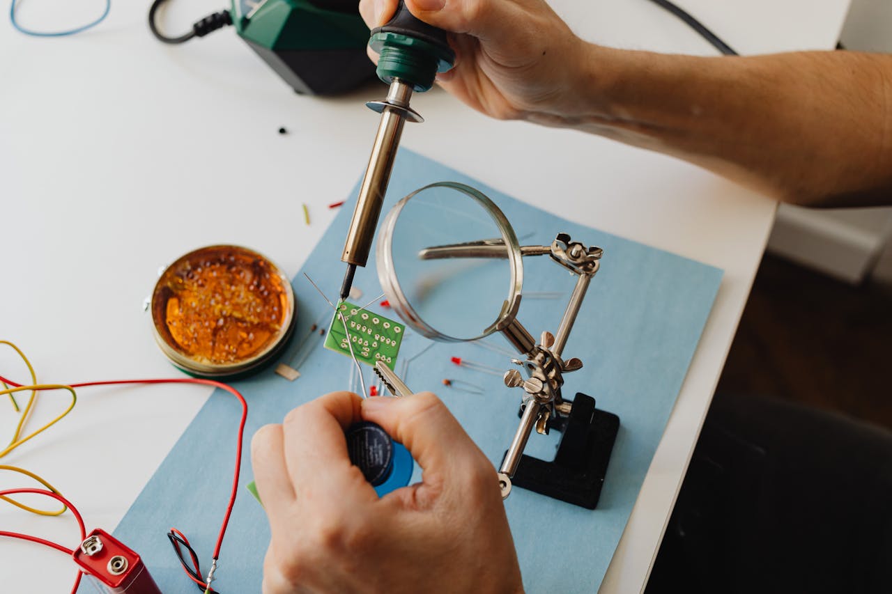 A detailed view of a technician using a soldering iron on a circuit board through a magnifying glass.